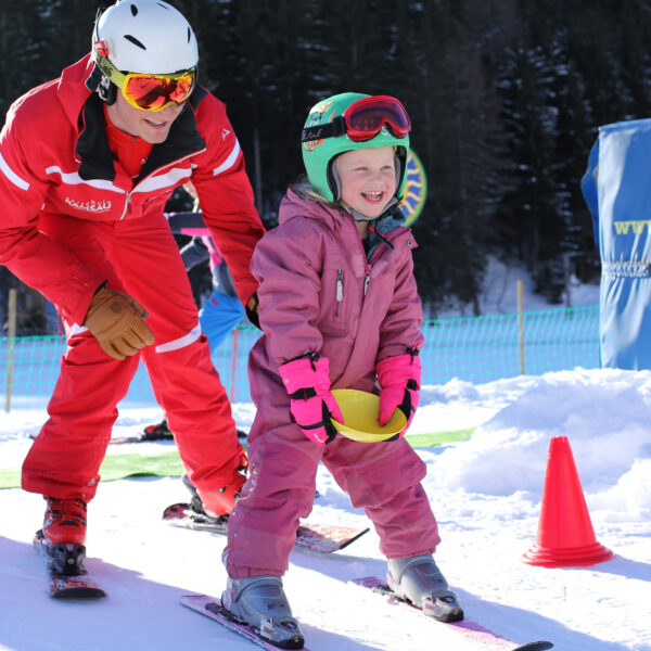 Ein erwachsener Skilehrer hilft einem lächelnden Kind in rosa Skikleidung auf einer verschneiten Piste, mit Sicherheitskegeln und schneebedeckten Bäumen im Hintergrund.