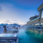 Ein beheizter Infinity-Pool im Freien eines modernen Hotels bietet in der Abenddämmerung einen Blick auf verschneite Dächer und ferne Berge. Dampf steigt aus dem Wasser in die kalte Abendluft auf.