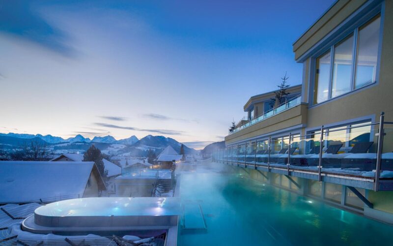Ein beheizter Infinity-Pool im Freien eines modernen Hotels bietet in der Abenddämmerung einen Blick auf verschneite Dächer und ferne Berge. Dampf steigt aus dem Wasser in die kalte Abendluft auf.