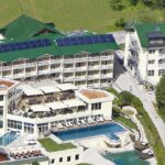 Luftaufnahme eines großen Hotelkomplexes mit mehreren Gebäuden mit begrünten Dächern, Außenpools, Sonnenterrassen und umliegenden Grünflächen.