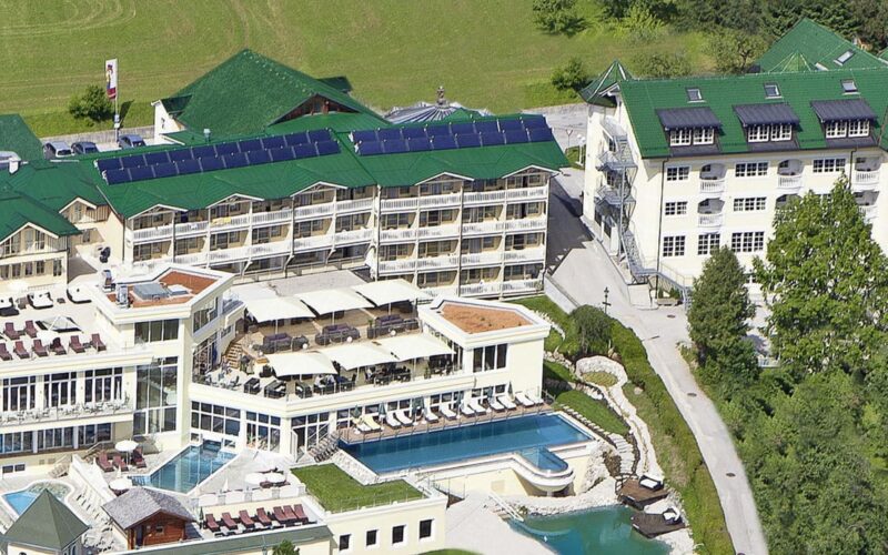 Luftaufnahme eines großen Hotelkomplexes mit mehreren Gebäuden mit begrünten Dächern, Außenpools, Sonnenterrassen und umliegenden Grünflächen.