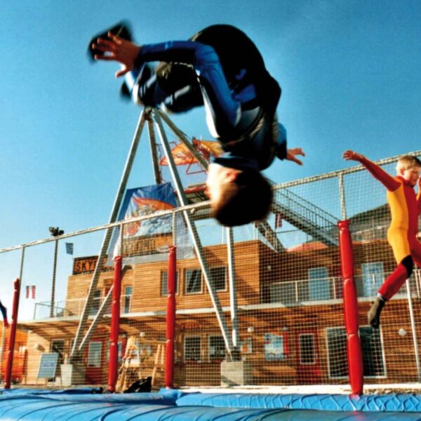 Zwei Kinder in Overalls hüpfen draußen auf einem Trampolin; eines macht gerade einen Salto, während das andere in der Luft ist. Im Hintergrund sind Gebäude und ein Zaun zu sehen.