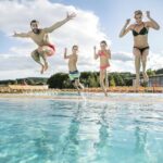 Vier Personen, zwei Erwachsene und zwei Kinder, springen an einem sonnigen Tag gemeinsam in ein Freibad.