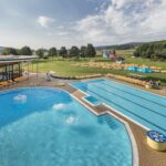 Freibadkomplex mit einem runden und einem rechteckigen Becken, vielen gelben Liegestühlen auf dem Rasen und einem Gebäude in der Nähe unter einem teilweise bewölkten Himmel.