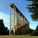 Eine hohe Holzkonstruktion mit Treppe steht unter freiem Himmel, umgeben von Bäumen, unter einem blauen Himmel.