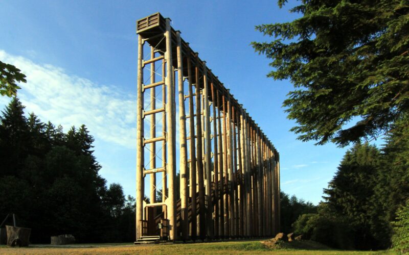 Eine hohe Holzkonstruktion mit Treppe steht unter freiem Himmel, umgeben von Bäumen, unter einem blauen Himmel.
