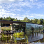 Ein modernes Glasgebäude mit Flachdach liegt neben einem Teich, umgeben von Bäumen und Gras, auf dessen Außenterrasse sich Menschen versammeln.