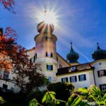 Das Sonnenlicht scheint direkt auf den Turm eines historischen weißen Gebäudes mit grünen Kuppeln, umgeben von Laub und unter einem klaren blauen Himmel.