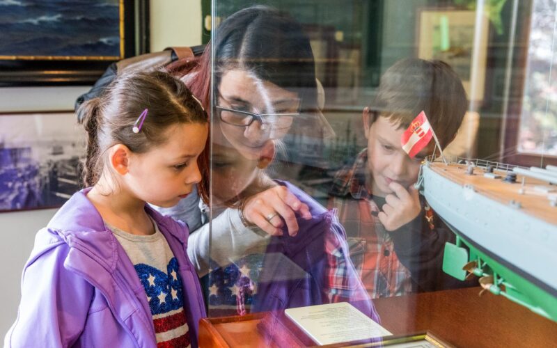Eine Frau und zwei Kinder sehen sich eine Museumsausstellung mit einem Schiffsmodell hinter Glas genau an und lesen eine Informationstafel.