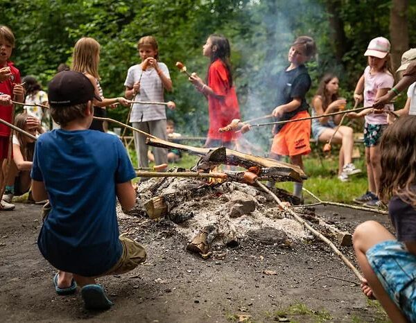 Kinder rösten Essen auf Stöcken über einem Lagerfeuer in einem bewaldeten Außenbereich, während Rauch aufsteigt und andere in der Nähe sitzen oder stehen.