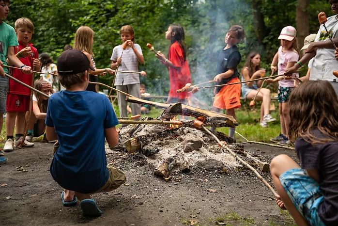 Kinder rösten Essen auf Stöcken über einem Lagerfeuer in einem bewaldeten Außenbereich, während Rauch aufsteigt und andere in der Nähe sitzen oder stehen.