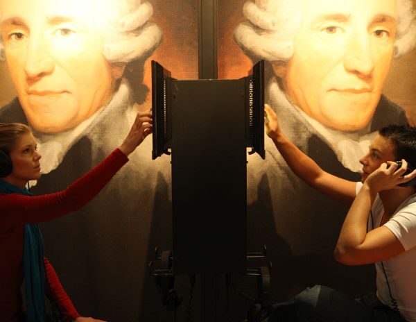 An einer Audiostation sitzen sich zwei Personen mit Kopfhörern gegenüber, im Hintergrund sind große Porträts von Franz Joseph Haydn zu sehen.