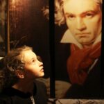 Ein kleines Kind blickt zu einem großen Porträt von Ludwig van Beethoven an der Wand eines Museums auf.