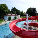 Eine große rote Wasserrutsche führt in einen Swimmingpool im Freien; an sonnigen Tagen entspannt man sich auf dem Rasen und den Liegestühlen unter den Bäumen.
