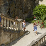 Mehrere Kinder laufen und rennen auf Steintreppen und einem Kiesweg in einem Gartenbereich mit grünen Sträuchern und blühenden rosa Blumen.