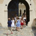 Eine Gruppe von Kindern geht gemeinsam durch ein bogenförmiges Tor in ein Gebäude mit Steinpflaster und cremefarbenen Wänden.