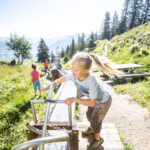 Ein junges Mädchen dreht an einem sonnigen Tag im Freien eine Wasserpumpe mit Metallspirale, während im grasbewachsenen, bergigen Hintergrund Kinder spielen.