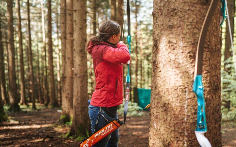 Eine Person in einer roten Jacke zielt mit Pfeil und Bogen in einem Waldstück, im Vordergrund lehnt ein weiterer Bogen an einem Baum.