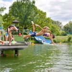 Vier Kinder springen von einem Steg in einen See, im Hintergrund sind grüne Bäume und eine Wasserrutsche zu sehen.