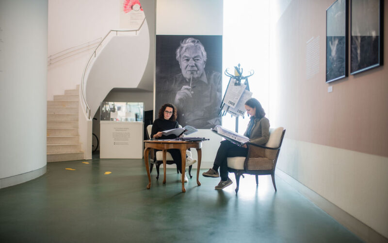 Zwei Personen sitzen an einem Tisch und lesen Dokumente in einem modernen Galerieraum, in dem ein großes Schwarz-Weiß-Porträt und Fotos an den Wänden hängen.