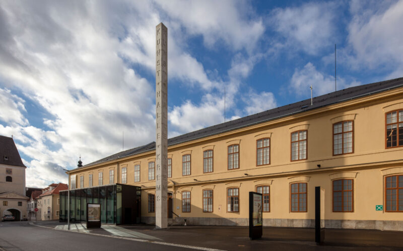 Ein großes beigefarbenes Gebäude mit gewölbten Fenstern und einem hohen vertikalen Schild mit der Aufschrift "KUNSTHALLE" unter einem teilweise bewölkten Himmel.