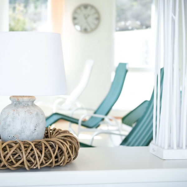 Eine weiße Lampe mit einem Korbsockel steht auf einer weißen Fläche; im Hintergrund stehen tealfarbene Loungesessel, eine Uhr und helle Fenster.