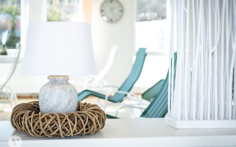 Eine weiße Lampe mit einem Korbsockel steht auf einer weißen Fläche; im Hintergrund stehen tealfarbene Loungesessel, eine Uhr und helle Fenster.