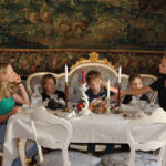 Fünf Kinder sitzen um einen eleganten Esstisch mit Tee und Essen in einem Raum, der mit einem kunstvollen Wandteppich und alten Möbeln geschmückt ist.