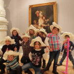 Eine Gruppe von Kindern mit großen Papierhüten posiert vor einem klassischen Gemälde in einem Kunstmuseum.