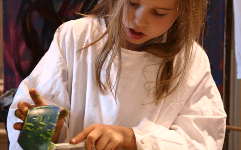 Ein junges Mädchen in einem weißen Kittel malt mit einem Pinsel an einem Tisch, umgeben von bunten Schalen und Kunstzubehör, mit abstrakten Kunstwerken im Hintergrund.