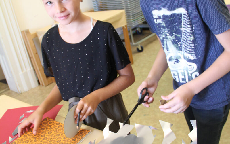 Zwei Kinder stehen an einem Tisch und schneiden mit einer Schere Dekorationspapier und Silberfolie aus, um im Haus zu basteln.