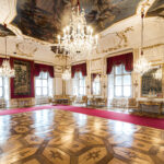 Opulenter Palastsaal mit kunstvollen Kronleuchtern, roten Vorhängen, Parkettböden, goldverzierten Wänden, großen Spiegeln und aufwändigen Deckengemälden.