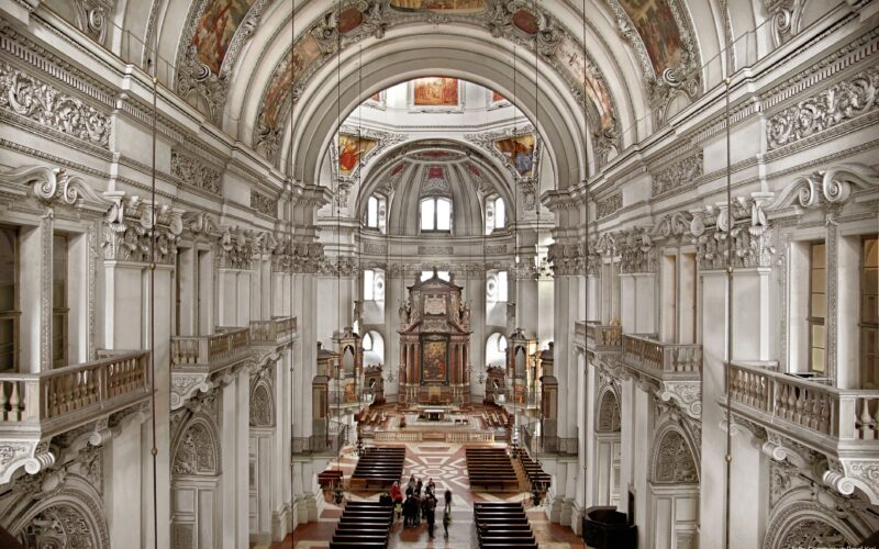 Barockes Kircheninnere mit verschnörkelten Bögen, Säulen, dekorativen Decken und einer Gruppe von Menschen, die sich vorne am Altar versammelt haben.