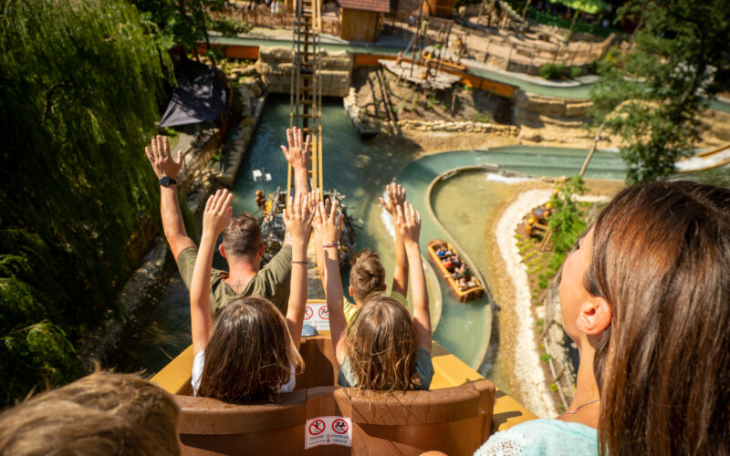 Eine Gruppe von Menschen mit erhobenen Händen fährt in einem Vergnügungspark eine Wasserrutsche steil hinunter, wobei das Wasser und die Parklandschaft unten sichtbar sind.