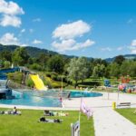 Freibadkomplex mit Wasserrutschen, umgeben von Grünflächen zum Sonnenbaden und Entspannen vor einer Kulisse aus grünen Hügeln und Bäumen unter blauem Himmel.