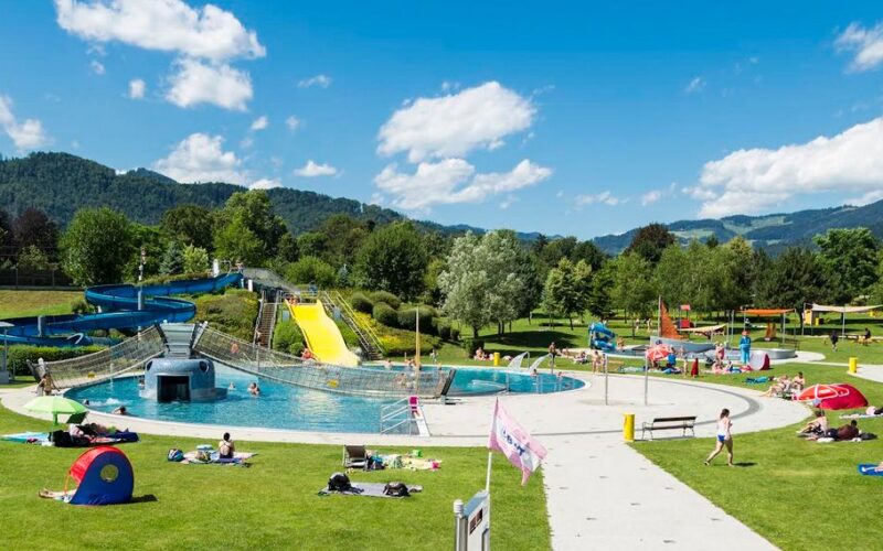 Freibadkomplex mit Wasserrutschen, umgeben von Grünflächen zum Sonnenbaden und Entspannen vor einer Kulisse aus grünen Hügeln und Bäumen unter blauem Himmel.