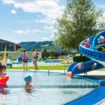 Kinder spielen in einem flachen Außenbecken neben einer blauen Schlangenrutsche, während Erwachsene an einem sonnigen Tag im Hintergrund zuschauen.