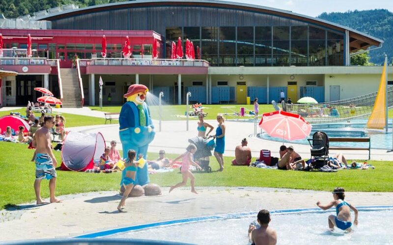 Familien und Kinder erfreuen sich an einem grasbewachsenen Außenbereich mit Pools, Zelten und einer großen Zeichentrickfiguren-Statue in der Nähe eines modernen Gebäudes mit großen Fenstern.
