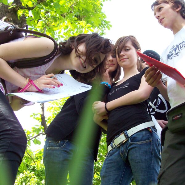 Eine Gruppe Teenager steht im Freien im Grünen, einige halten Papier und Stifte in den Händen und scheinen an einer Outdoor-Aktivität oder einem Studium teilzunehmen.