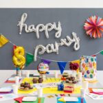 Ein geschmückter Geburtstagstisch mit bunten Tassen, Tellern, Luftballons, Muffins, einer Torte, Geschenken und einem "Happy Party"-Ballonbanner an der Wand.
