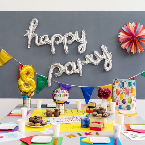 Ein geschmückter Geburtstagstisch mit bunten Tassen, Tellern, Luftballons, Muffins, einer Torte, Geschenken und einem "Happy Party"-Ballonbanner an der Wand.