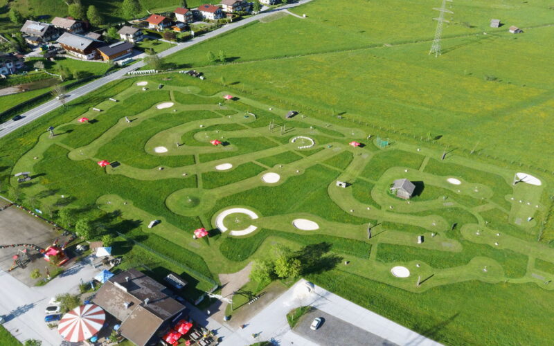 Luftaufnahme eines Minigolfplatzes mit gewundenen Wegen, kreisförmigen Grüns, Hindernissen und roten Sonnenschirmen, umgeben von Grasfeldern und nahe gelegenen Gebäuden.