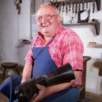 Ein älterer Mann mit Brille und blauer Schürze hält einen großen schwarzen Stiefel in der Hand. Er sitzt in einer Werkstatt, hinter ihm hängen Werkzeuge an der Wand.