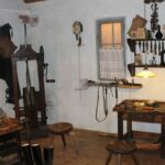 Eine alte Schusterwerkstatt mit Holzhockern, Werkzeugen an den Wänden, einer Nähmaschine, Schuhen und Werkbänken, beleuchtet von einer Hängelampe.