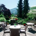 Terrasse mit Korbstühlen und Tischen auf einer Holzterrasse, umgeben von üppigem Grün und der Berglandschaft im Hintergrund.