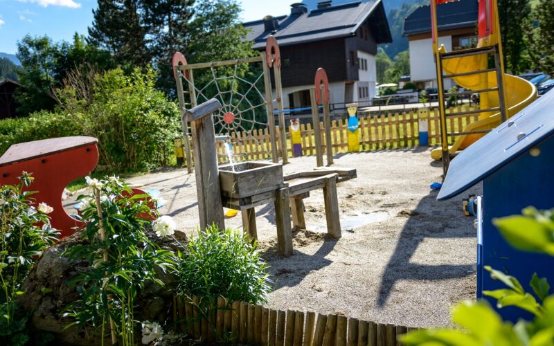 Spielplatz im Freien mit hölzernen Klettergerüsten, Rutsche, Bänken und Spielgeräten auf Sand, umgeben von Grünflächen und Häusern im Hintergrund.