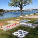 Ein großes Brettspiel im Freien, gemalt auf Gras in der Nähe eines Sees, mit farbigen Kreisen und Wegen, neben einem Baum und einem gepflasterten Gehweg.