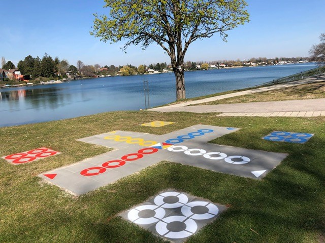 Ein großes Brettspiel im Freien, gemalt auf Gras in der Nähe eines Sees, mit farbigen Kreisen und Wegen, neben einem Baum und einem gepflasterten Gehweg.