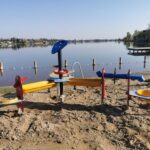 Ein Sand- und Wasserspielplatz für Kinder an einem sandigen Seeufer mit ruhigem Wasser, einem Steg und Häusern im Hintergrund.