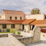 Steinhof mit Topfpflanzen und Fässern, umgeben von Gebäuden mit roten Ziegeldächern unter einem teilweise bewölkten Himmel.